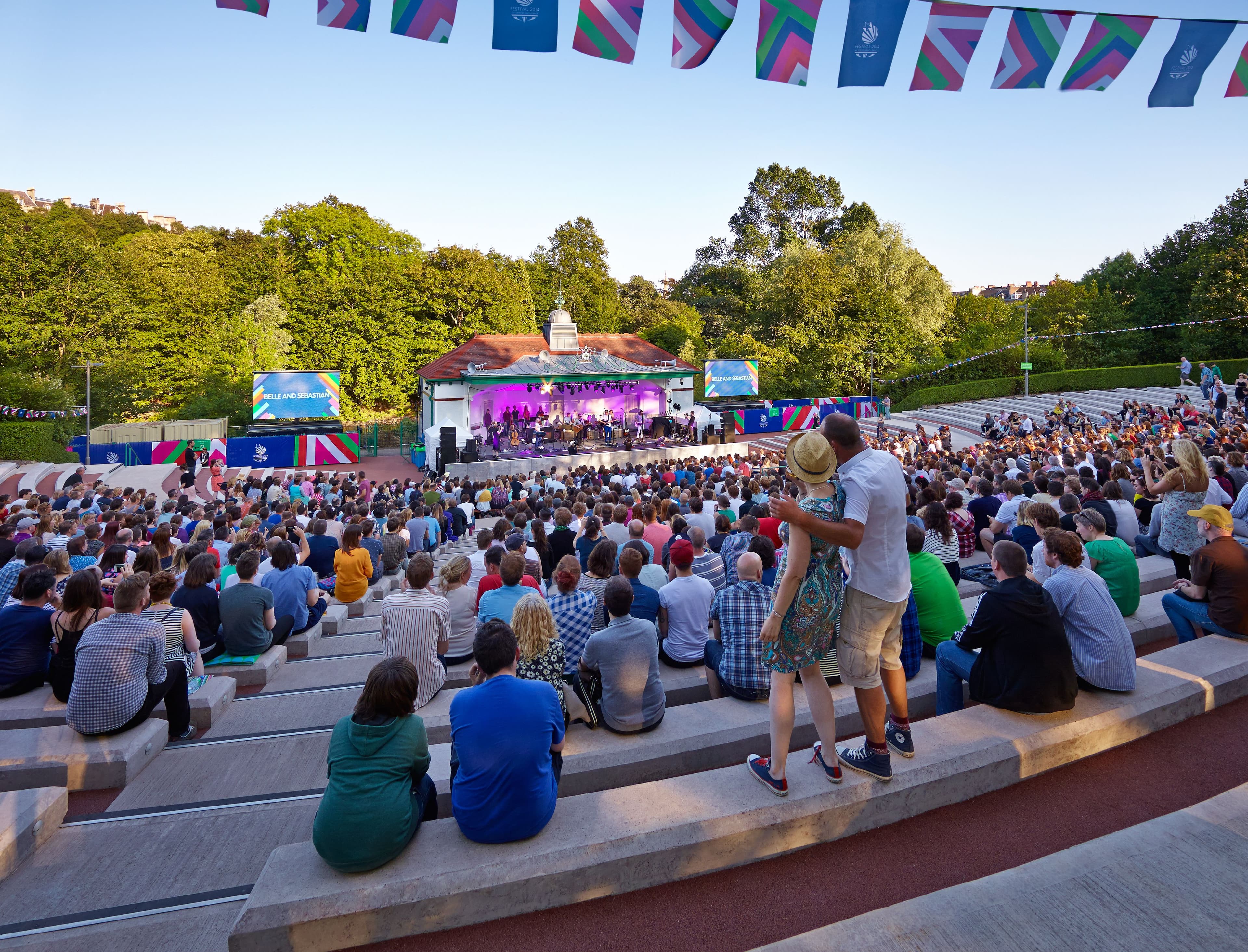 A photograph taken from the stands looking down towards the Kelvingrove Bandstand. People sit on the benches and bunting hangs above them.