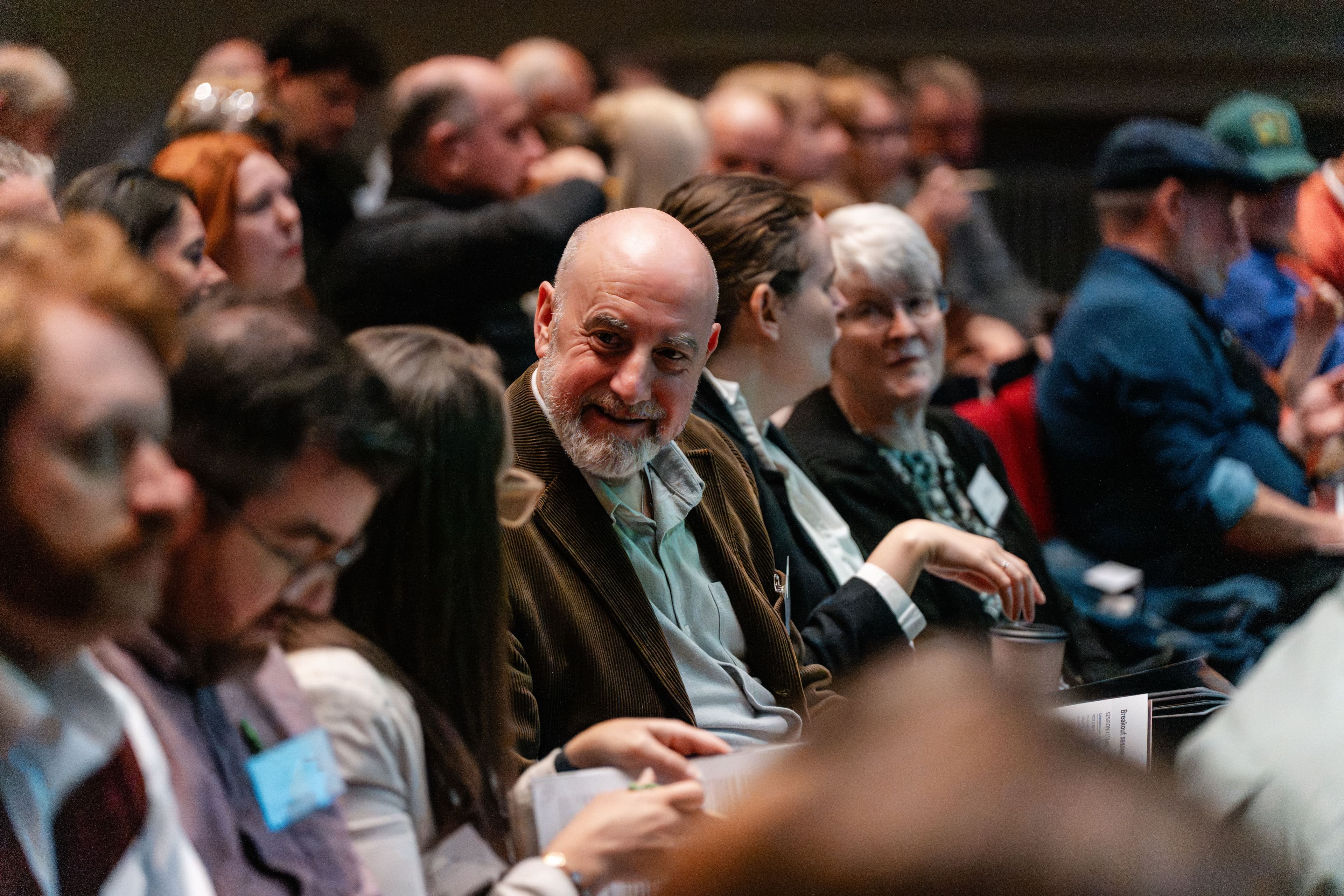 A photograph of rows of people sit in an auditorium. They are variously networking or looking ahead towards a stage.